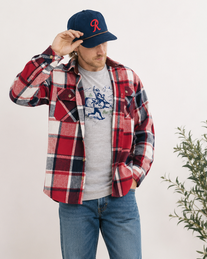 A man wearing a red and navy plaid flannel shirt over a gray graphic t-shirt, blue jeans, and brown suede shoes, standing in a room with a potted plant and a small wooden stool.