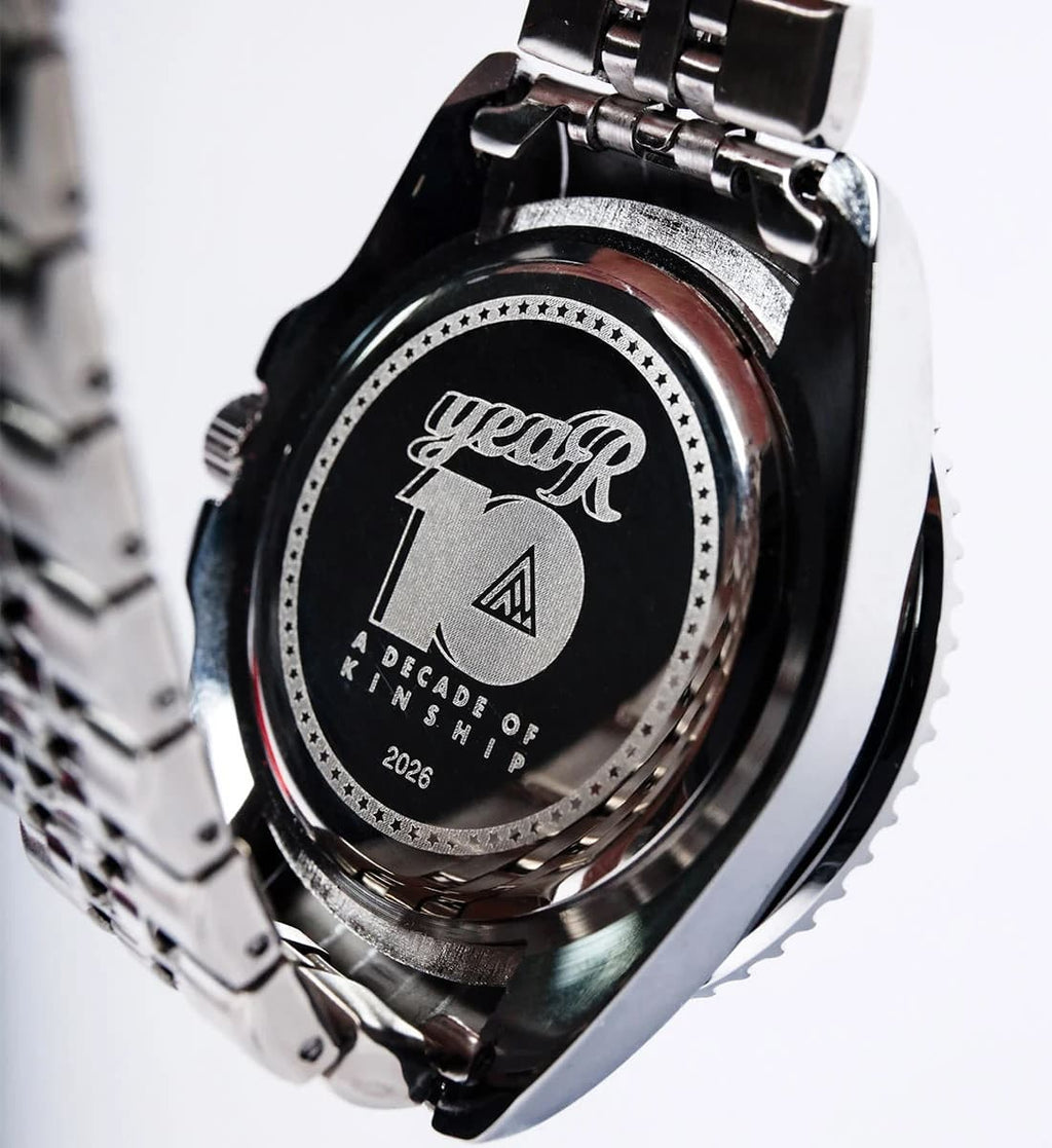 Close-up of the back of a stainless steel wristwatch with a polished metal bracelet, featuring an engraved black circular plate that reads 'year 10 A DECADE OF KINSHIP 2026'.