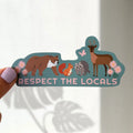 A hand with lavender nail polish holds a die-cut sticker against a white wall. The sticker features cartoon illustrations of a brown bear, red squirrel, gray raccoon, and brown deer on a teal background, with the text 'RESPECT THE LOCALS' in pink.