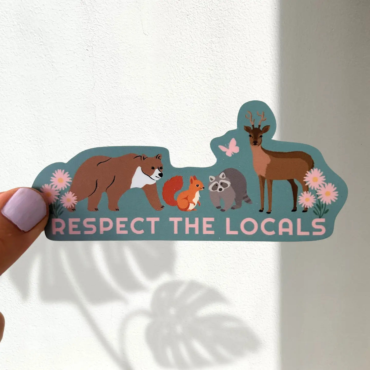 A hand with lavender nail polish holds a die-cut sticker against a white wall. The sticker features cartoon illustrations of a brown bear, red squirrel, gray raccoon, and brown deer on a teal background, with the text 'RESPECT THE LOCALS' in pink.