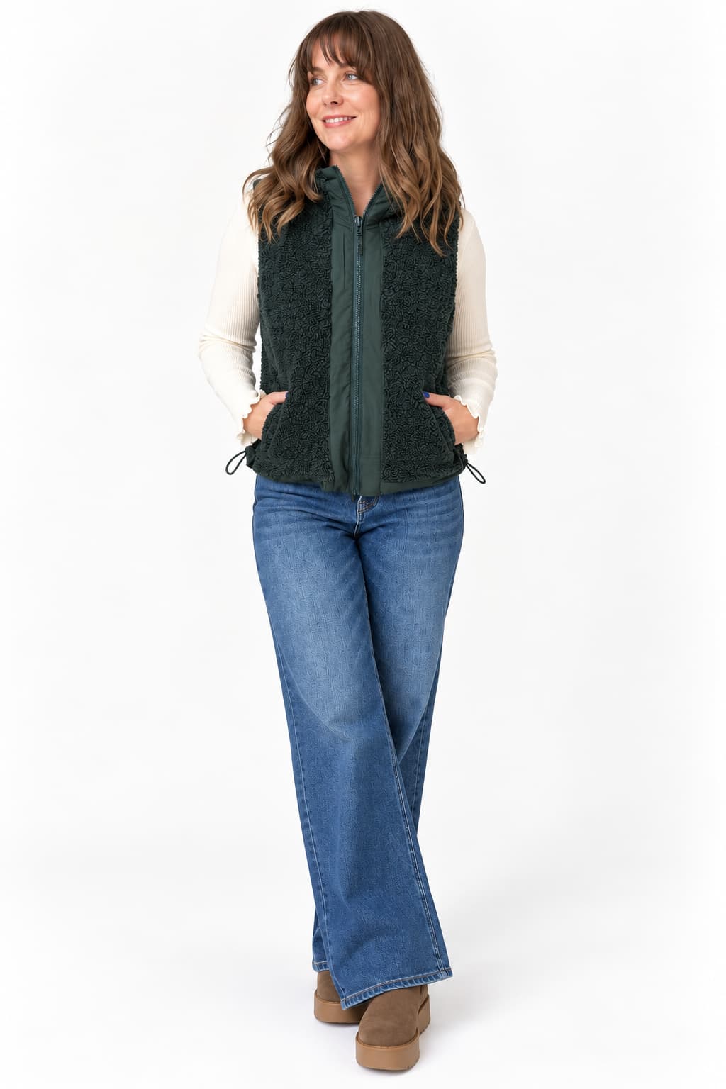 A woman modeling a dark green sherpa fleece vest over a cream long-sleeve shirt and blue jeans, standing against a plain white studio background.