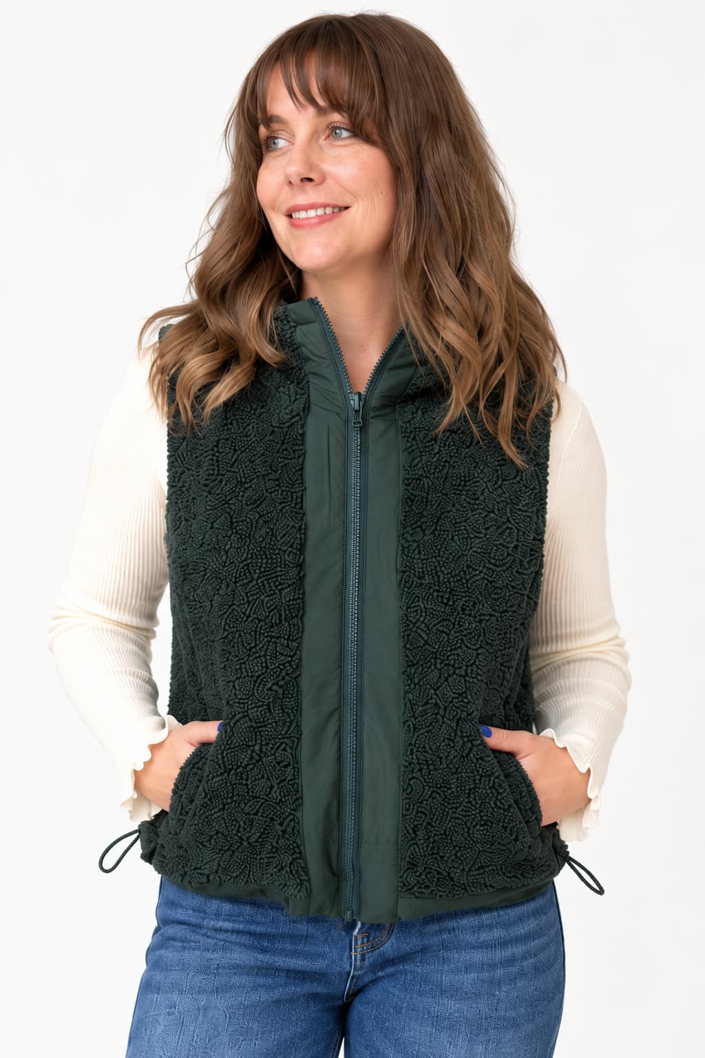 A woman wearing a dark forest green sherpa fleece zip-up vest over a cream ribbed long-sleeve shirt and blue jeans, standing against a plain white studio background.