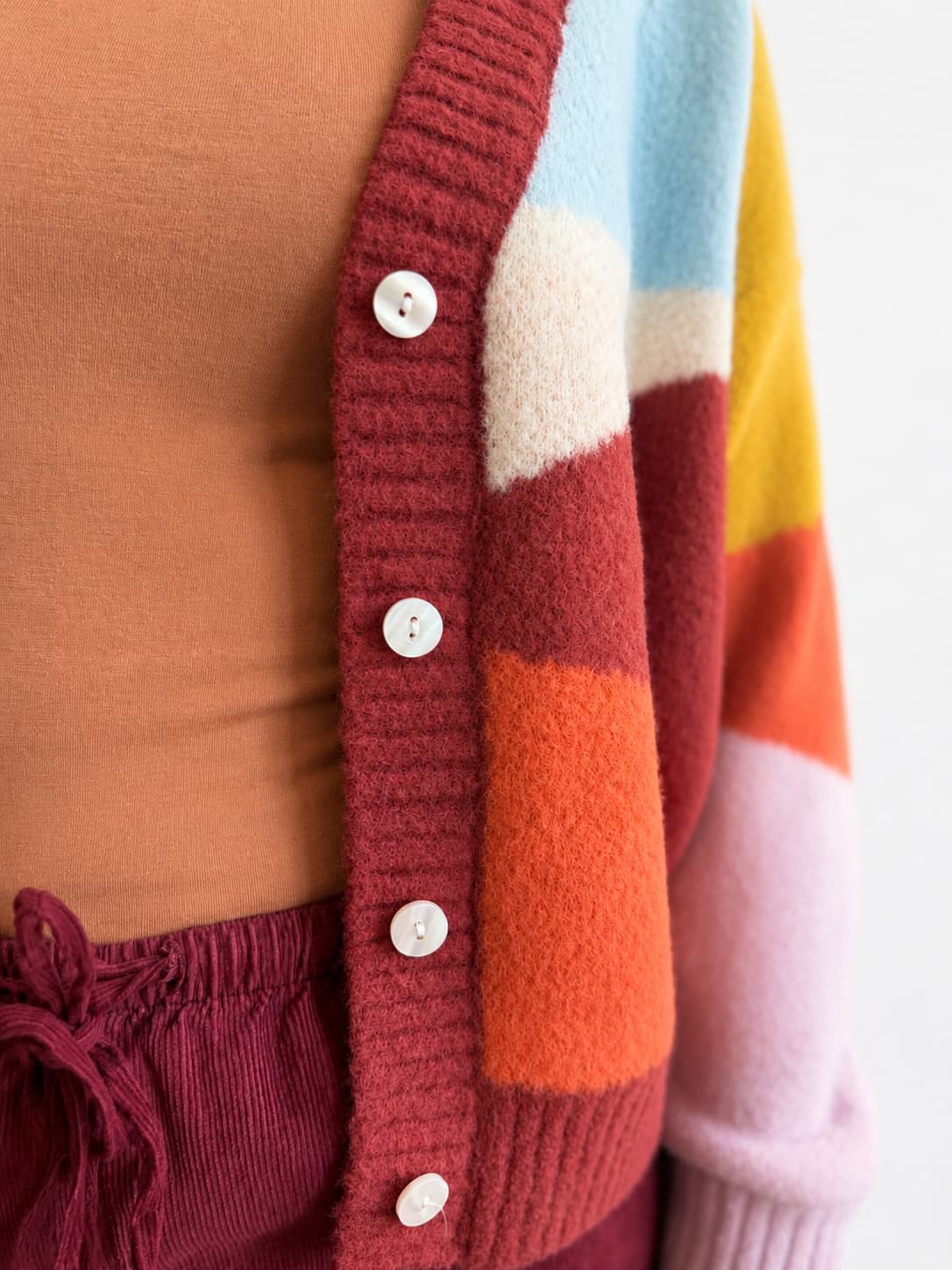 Close-up of a person wearing a chunky knit cardigan with large color-blocked panels in burgundy, orange, cream, light blue, yellow, and pale pink, over a rust-brown top and burgundy drawstring bottoms.