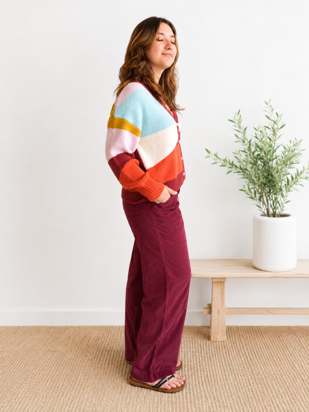 A woman stands in a bright room wearing a colorful striped knit sweater and burgundy wide-leg pants, with a small potted plant and light wood bench in the background.