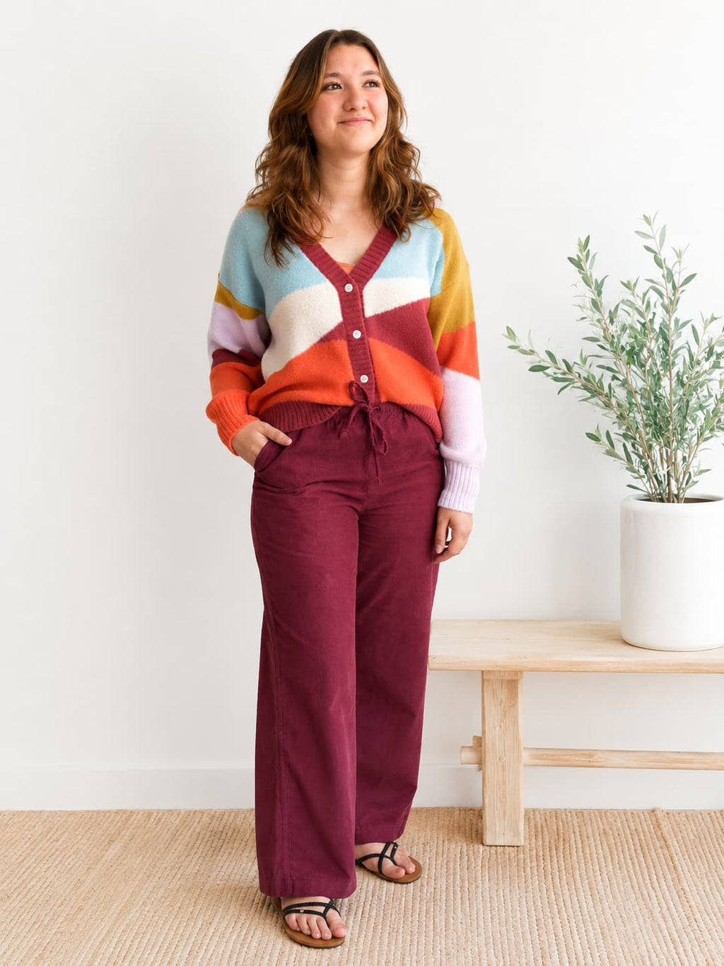 A woman modeling a colorful striped cardigan and burgundy wide-leg pants, standing in a bright room with a wooden bench and a potted plant.