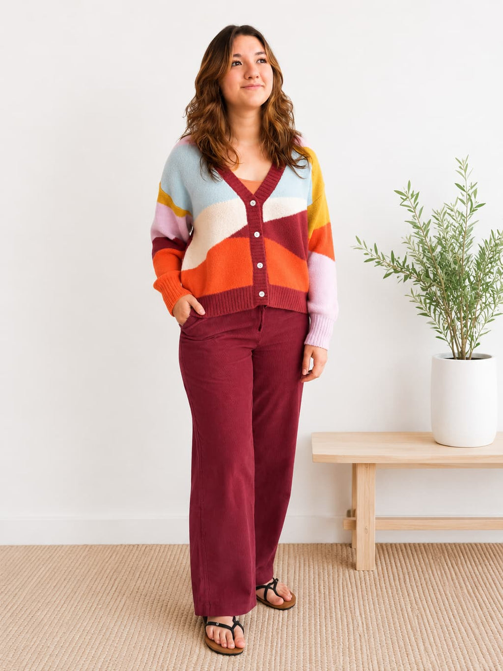 A woman stands in a bright room wearing a colorful cardigan with geometric color-block panels in light blue, cream, orange, burgundy, and pale pink, paired with burgundy wide-leg pants and sandals.