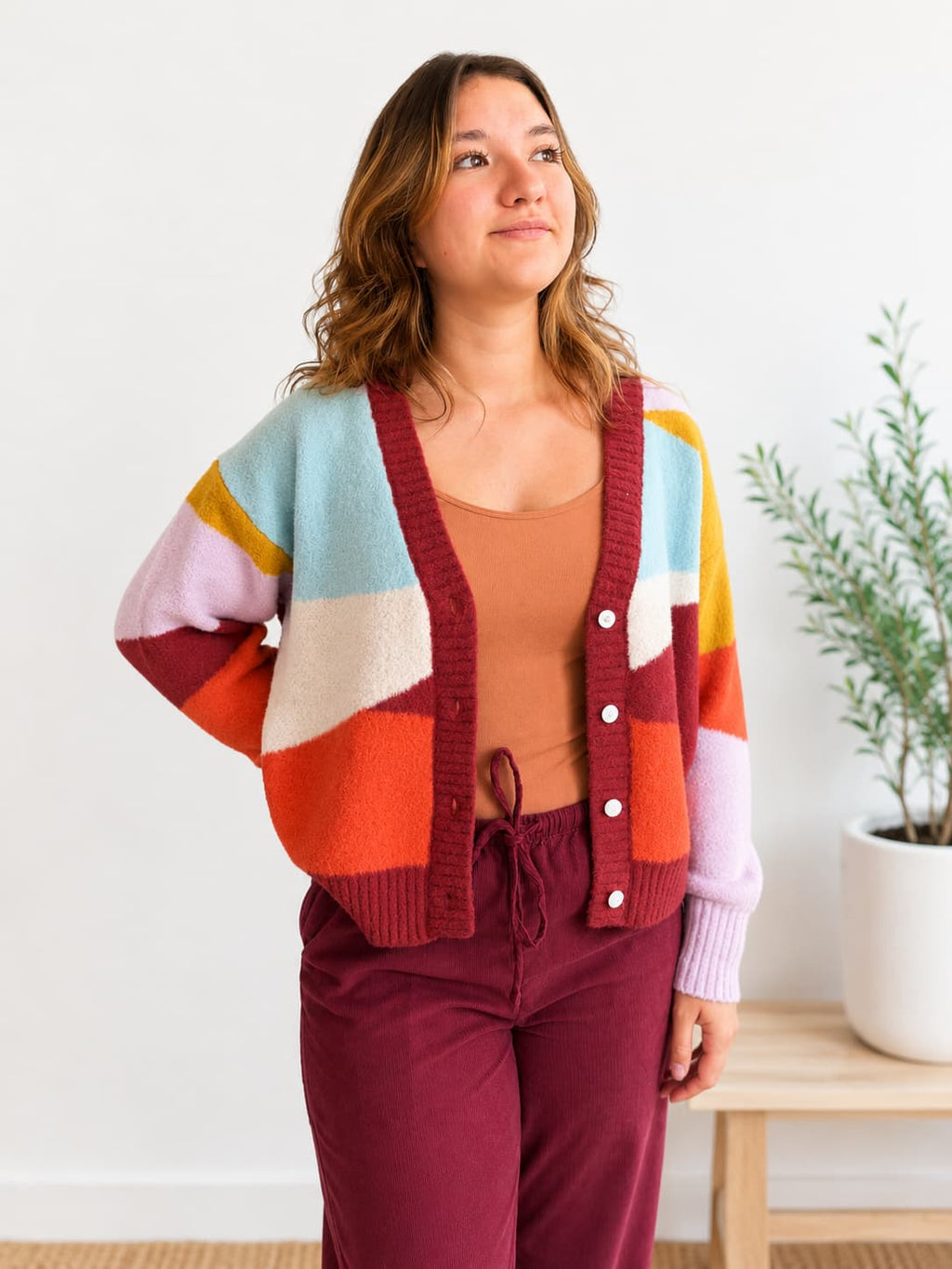 A woman wearing a colorful patchwork knit cardigan with red trim, styled with a rust-colored tank top and burgundy drawstring pants, standing in a bright indoor setting.