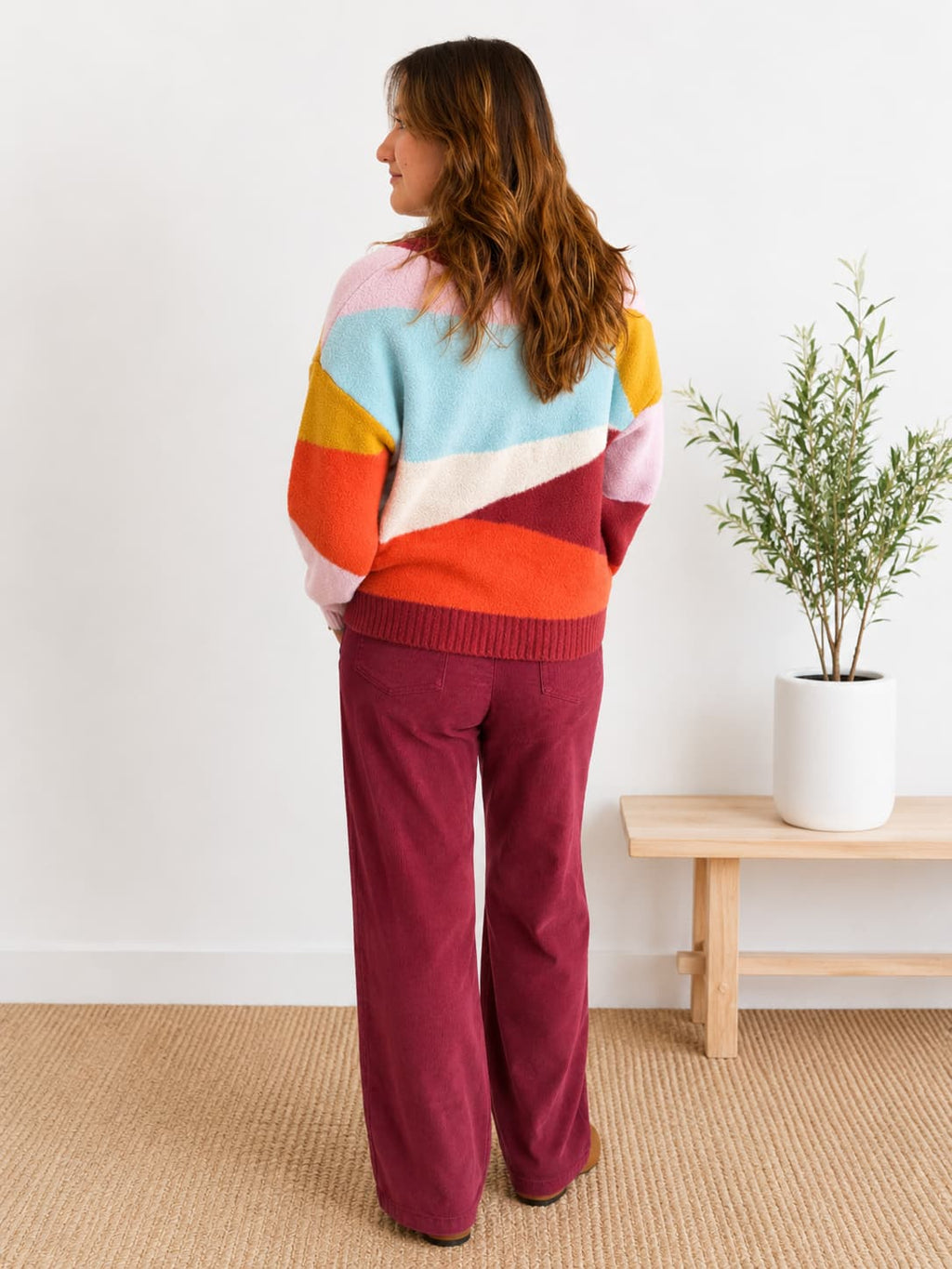 A woman stands with her back to the camera wearing a colorful striped knit sweater and burgundy wide-leg pants in a bright room with a small wooden bench and potted plant.