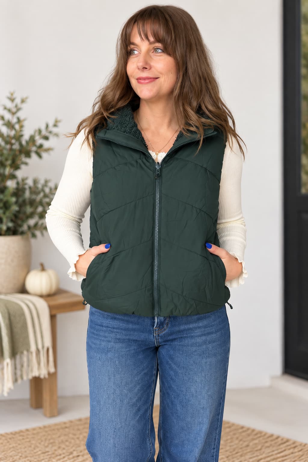 A woman wearing a dark forest green sleeveless puffer vest over a cream long-sleeve shirt and blue jeans, standing in a brightly lit indoor setting.