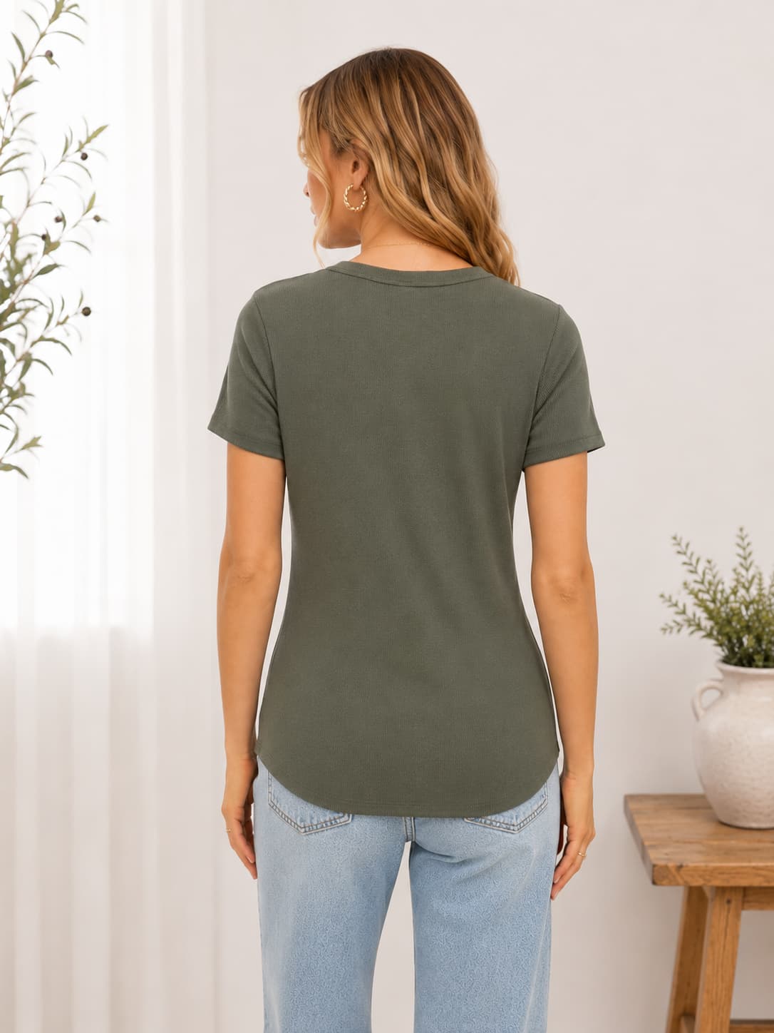 Woman wearing a ribbed green t-shirt and light blue jeans in a room with a plant and wooden table.