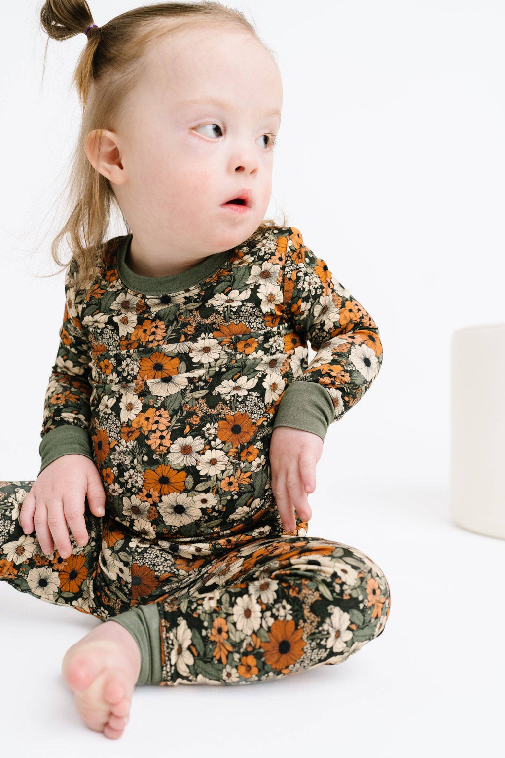 A toddler girl sitting cross-legged on a white floor, wearing a long-sleeve two-piece pajama set with an olive green base and a dense floral print in orange, cream, and brown.