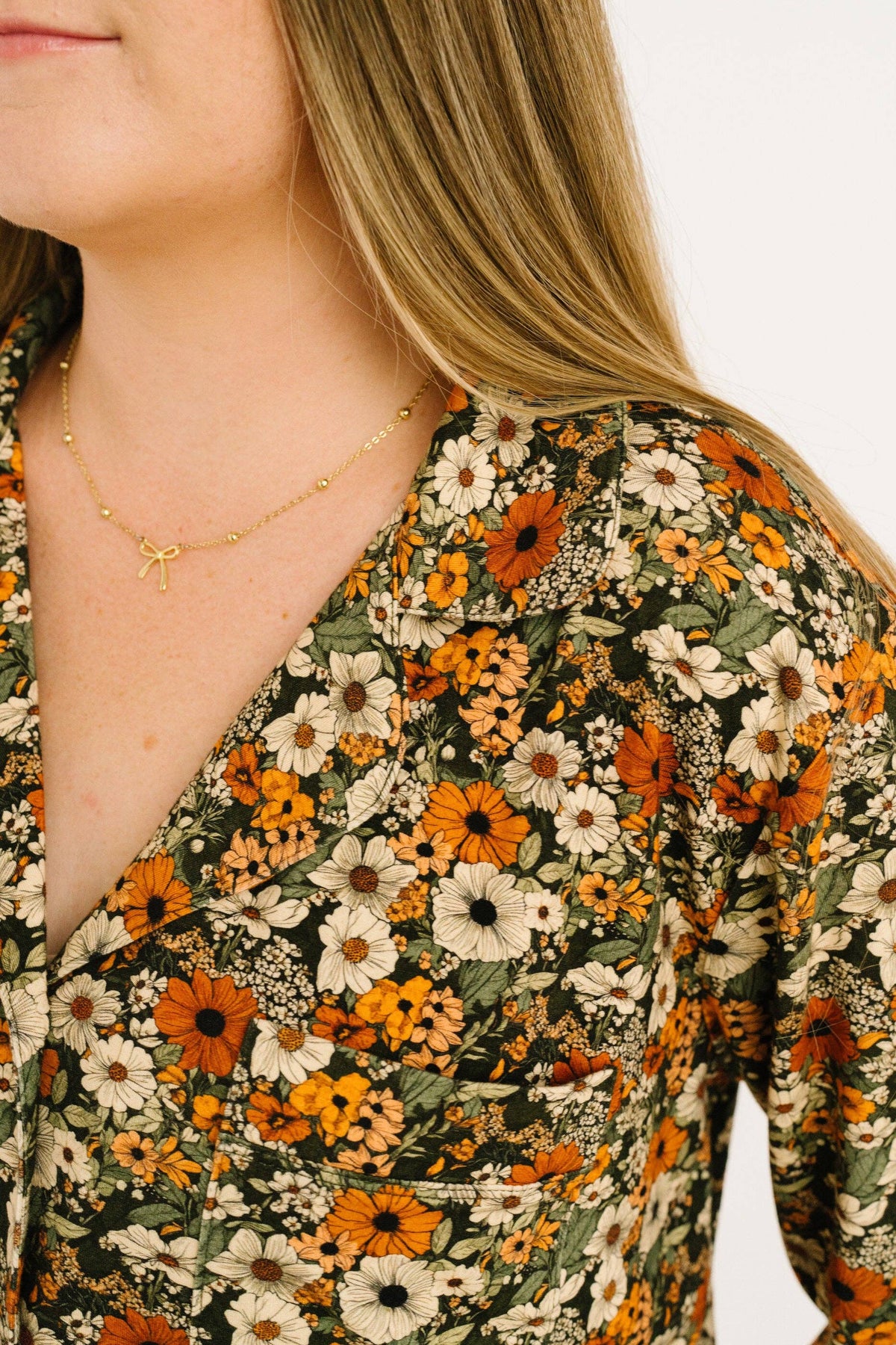 Close-up of a woman wearing a floral wrap-style top with a gold necklace. The top features an all-over print of orange, white, and green flowers on a dark background.