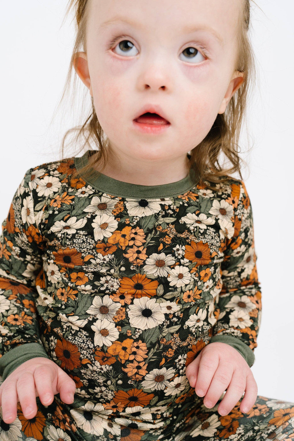 A close-up of a toddler wearing a long-sleeve pajama top with a dense floral print in orange, cream, and olive green on a dark background, set against a plain white studio background.