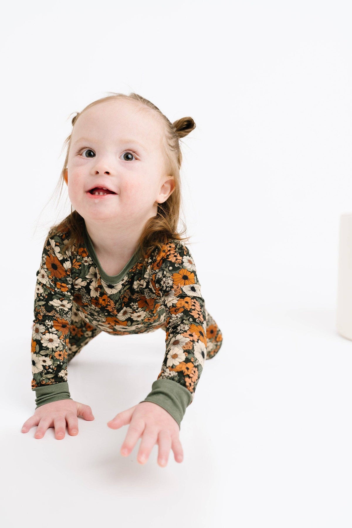 A toddler girl crawling on a white studio floor, wearing a long-sleeve pajama top with an orange, brown, and beige floral pattern and olive green cuffs.