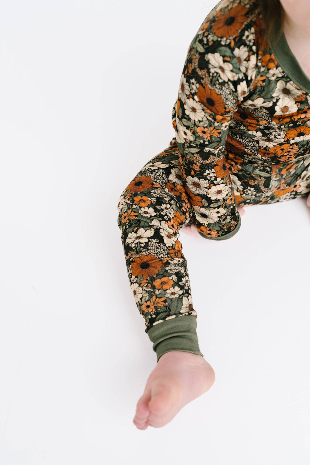 A close-up of a child wearing a long-sleeve pajama top with a dense floral print in orange, cream, and olive green, with a solid olive green crew neck and cuffs, shown against a plain white background.