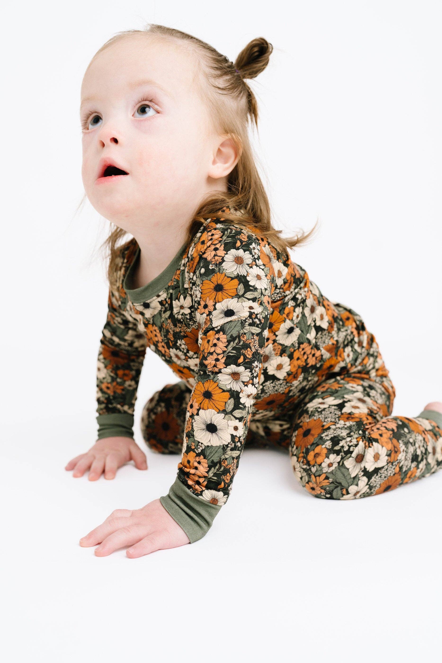 A toddler girl wearing a long-sleeve floral pajama set crawls on a white studio floor, looking upward with a surprised expression. The pajamas have an all-over floral print in orange, cream, and olive green, with solid olive green ribbed cuffs and collar.