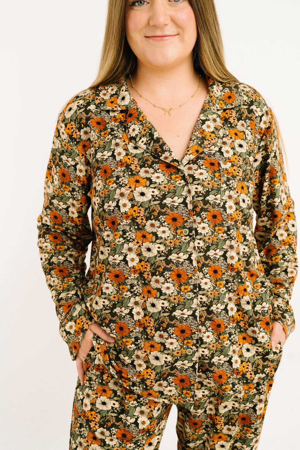 A woman wearing a long-sleeve button-front pajama top and matching pants in an olive green base fabric with a dense orange, yellow, and white floral print, photographed against a plain white background.
