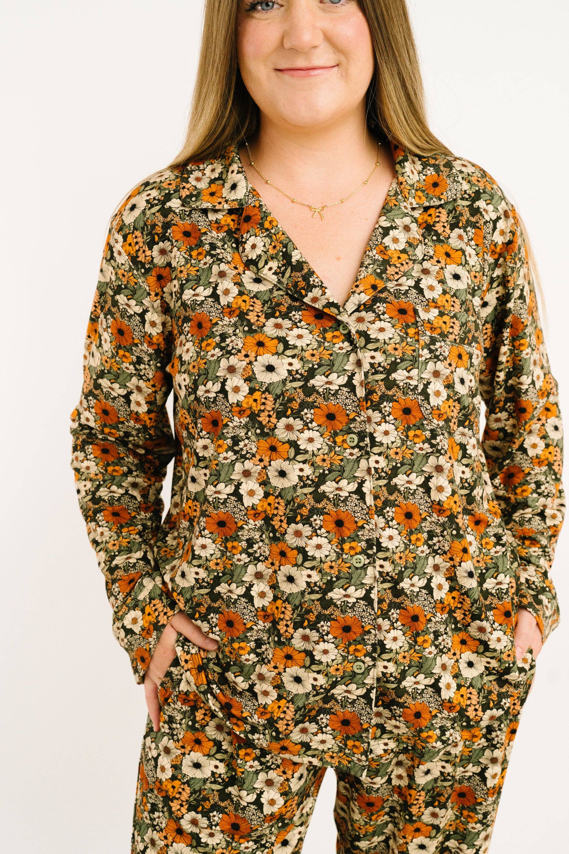 A woman wearing a long-sleeve button-front pajama top and matching pants in an olive green base fabric with a dense orange, yellow, and white floral print, photographed against a plain white background.