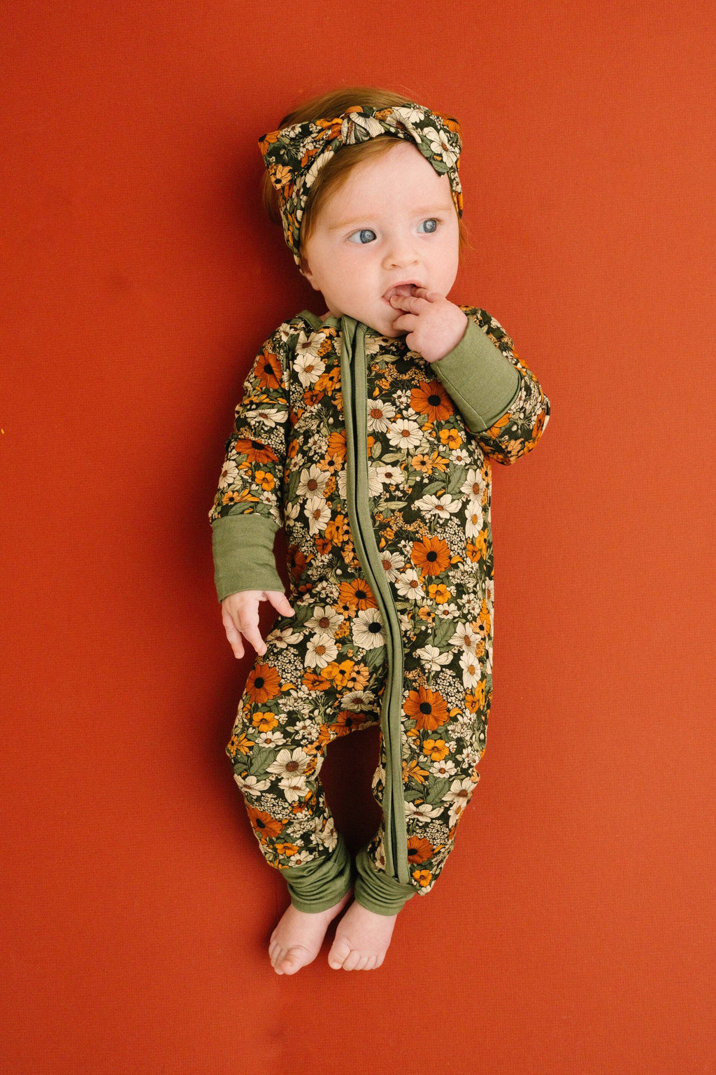 A baby wearing a floral print zip-up footless one-piece pajama with matching headband, standing against a solid terracotta orange background.