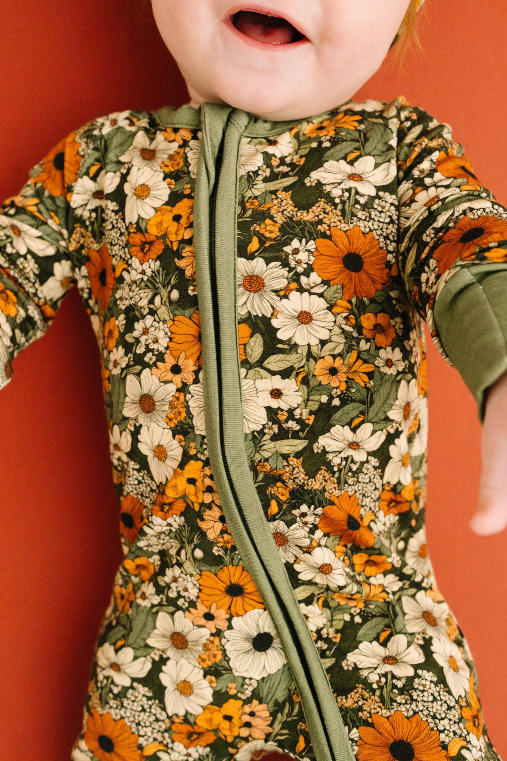 A close-up of a baby wearing a long-sleeve floral zip-up romper. The romper features a green base with a dense pattern of orange sunflowers, white daisies, and small wildflowers, accented by a solid sage green zipper and matching green cuffs.
