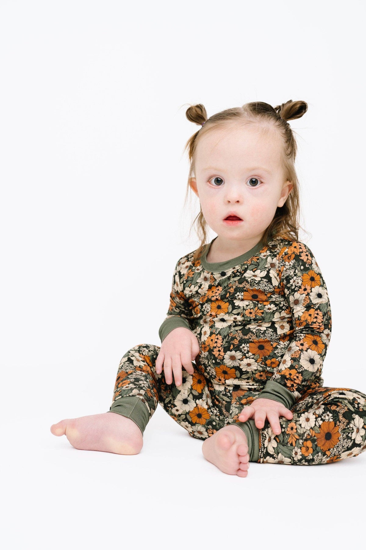 A toddler girl with her hair in two buns sits cross-legged on a white studio background, wearing a long-sleeve one-piece pajama set with an olive green base and a floral sunflower pattern in orange, brown, and cream.