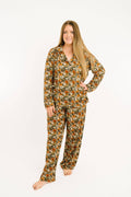 A woman modeling a two-piece long-sleeve pajama set with a dense floral print in orange, yellow, white, and green on a dark background, photographed against a solid white studio background.