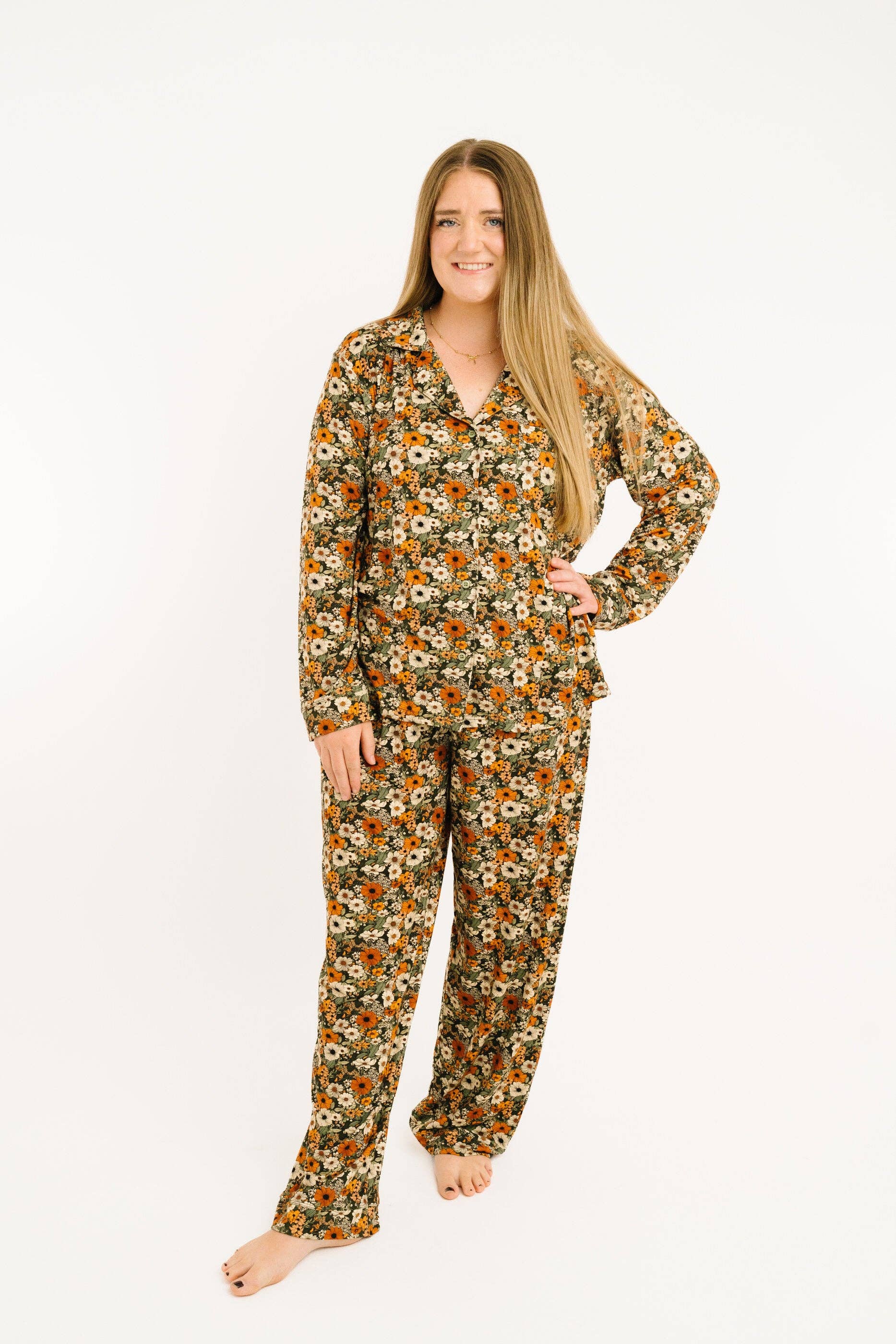 A woman modeling a two-piece long-sleeve pajama set with a dense floral print in orange, yellow, white, and green on a dark background, photographed against a solid white studio background.