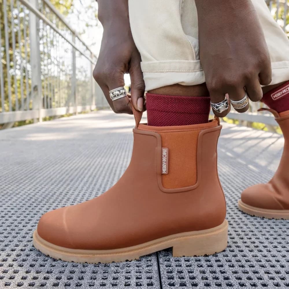 A person wearing rust Chelsea-style rain boots with elastic side panels and light beige soles, standing on a metal grate walkway outdoors. The person is pulling the boot up with both hands, which are adorned with silver rings, and a burgundy sock is visible above the boot.