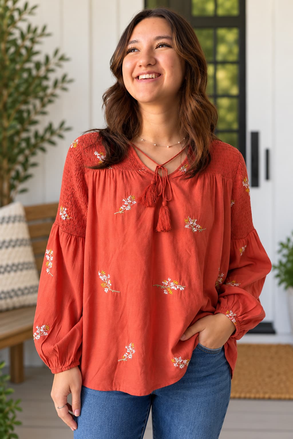 A woman wearing a rust-orange long-sleeve blouse with white floral embroidery and a tie-neck detail, paired with blue jeans, standing in a bright, modern porch setting.