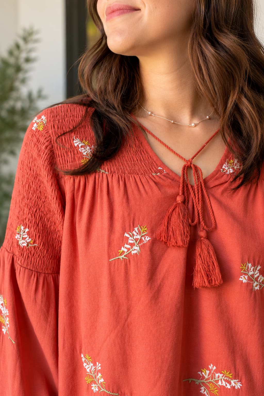 A close-up of a woman wearing a terracotta orange long-sleeve blouse with white floral embroidery and a tassel tie neckline.