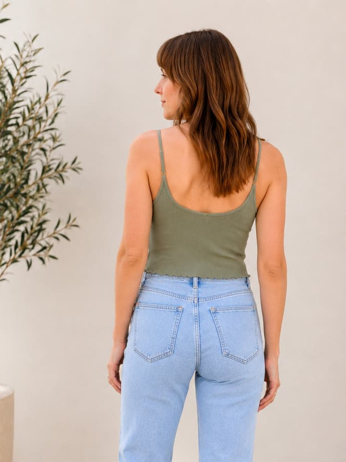 Woman wearing light blue jeans and a green tank top standing in a room with a plant and rug.