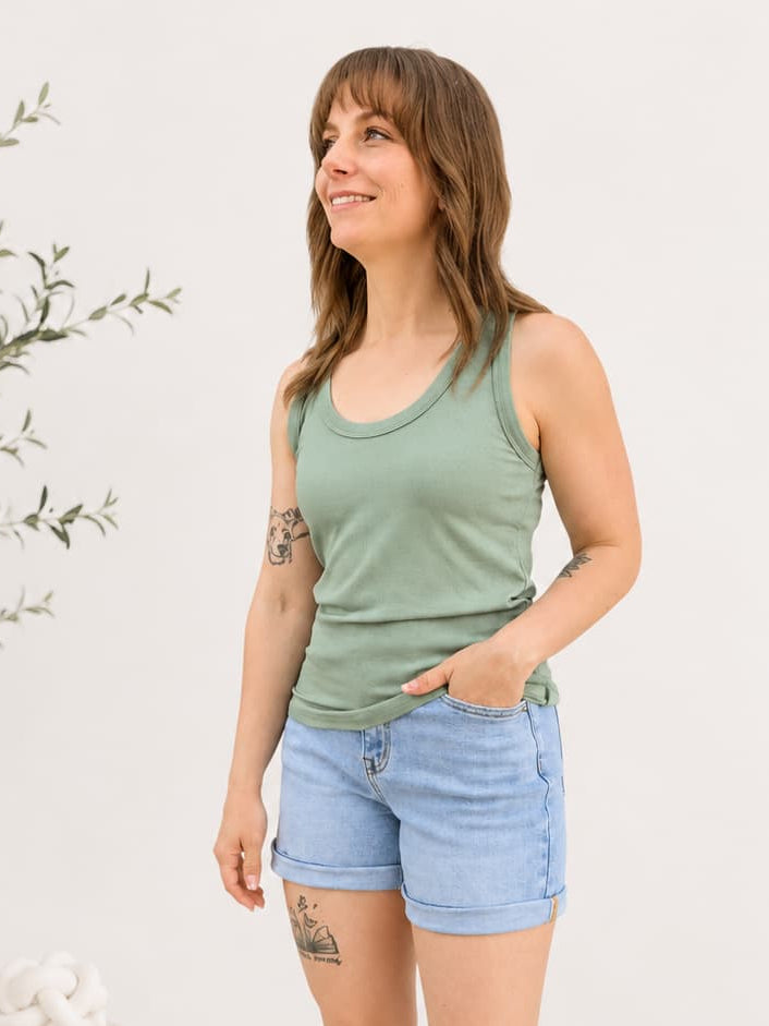 Woman wearing a green tank top and light blue shorts standing in a minimalistic room with a plant and white decor.