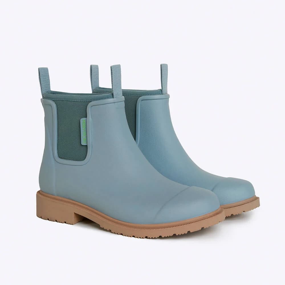 A pair of Chelsea-style rain boots in a dusty light blue color, featuring a contrasting dark green elastic side panel and a tan rubber sole, shown against a plain white background.