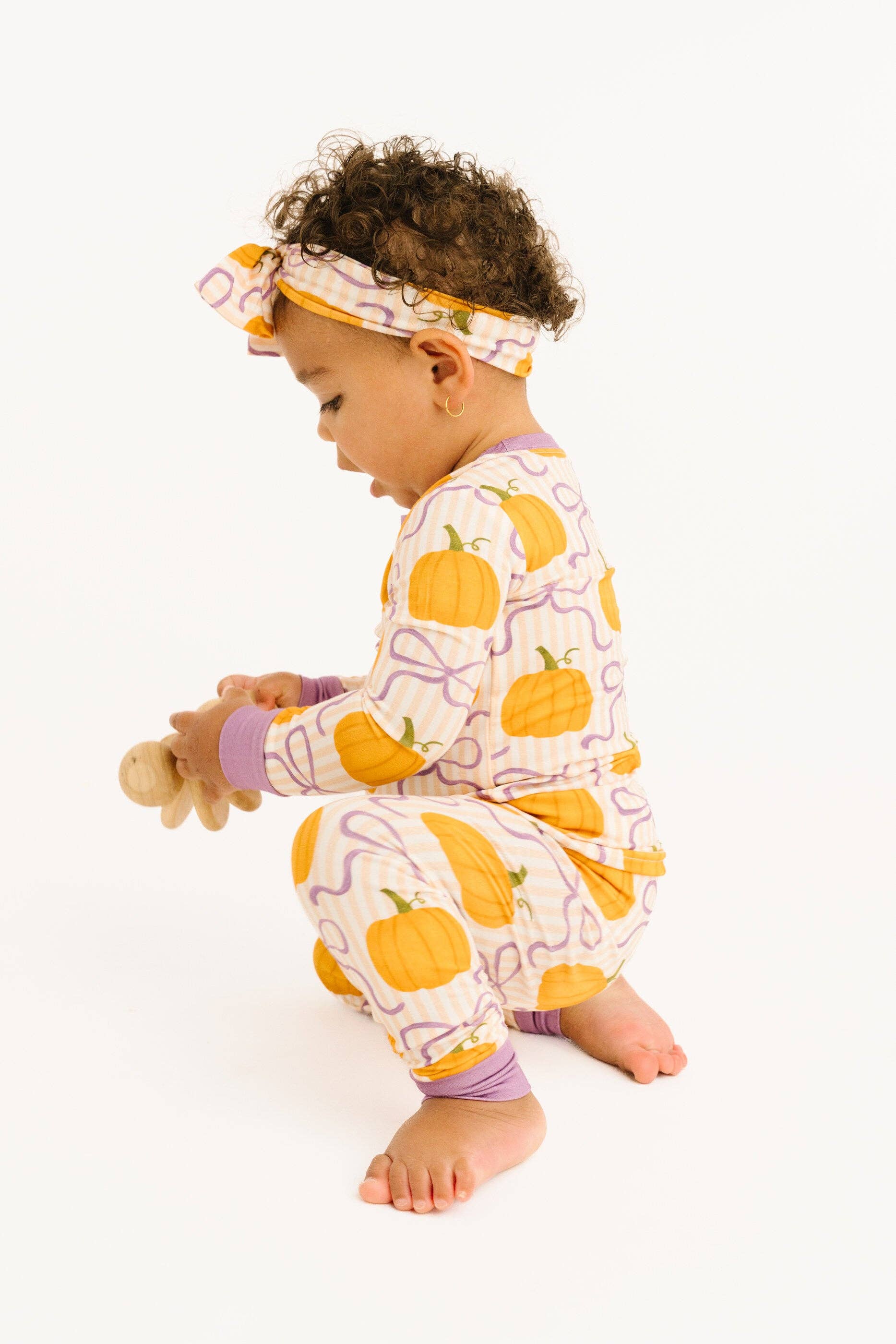 A toddler wearing a matching pajama set and headband with a pumpkin print, crouching on a white studio background while holding a wooden toy.