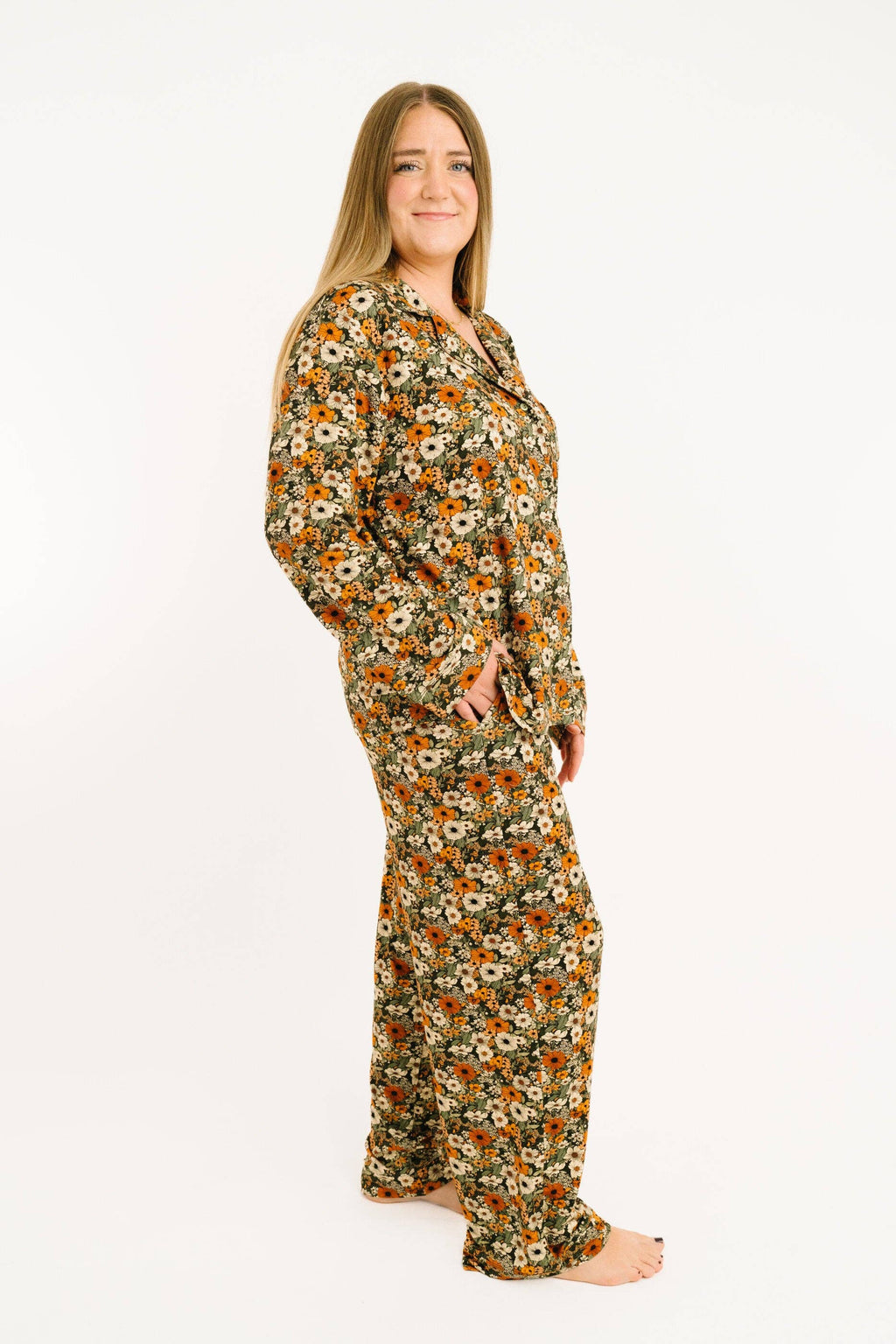 A woman modeling a long-sleeve pajama set with a dense floral print in orange, yellow, white, and green on a light background, standing against a plain white studio backdrop.