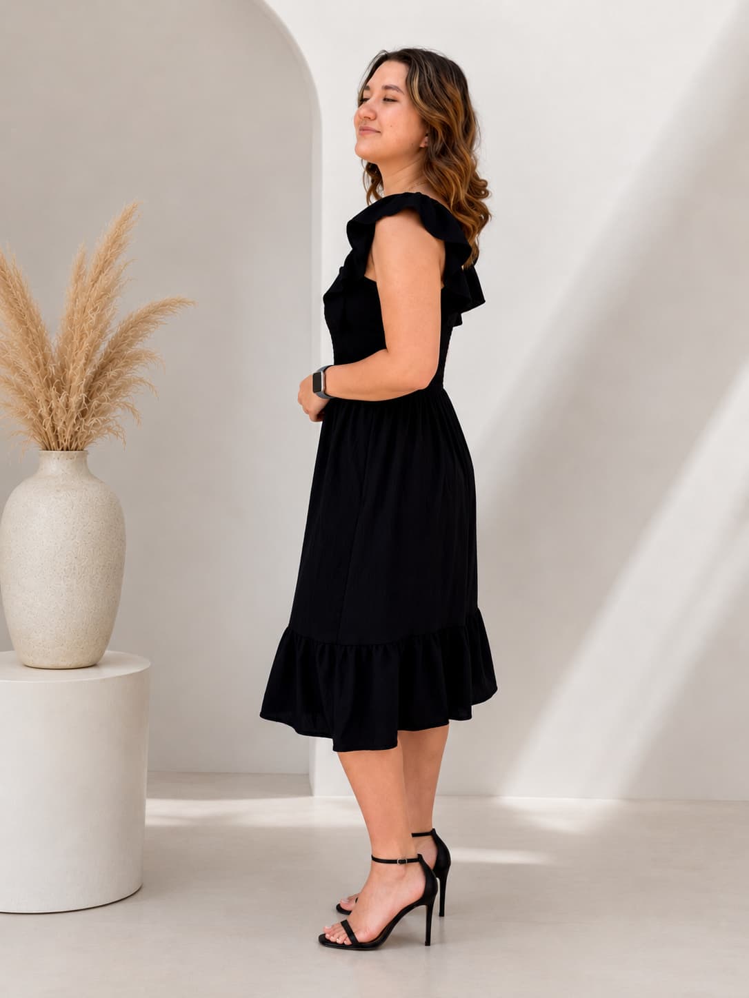 Woman wearing a black dress standing in a minimalistic room with a vase and plant.