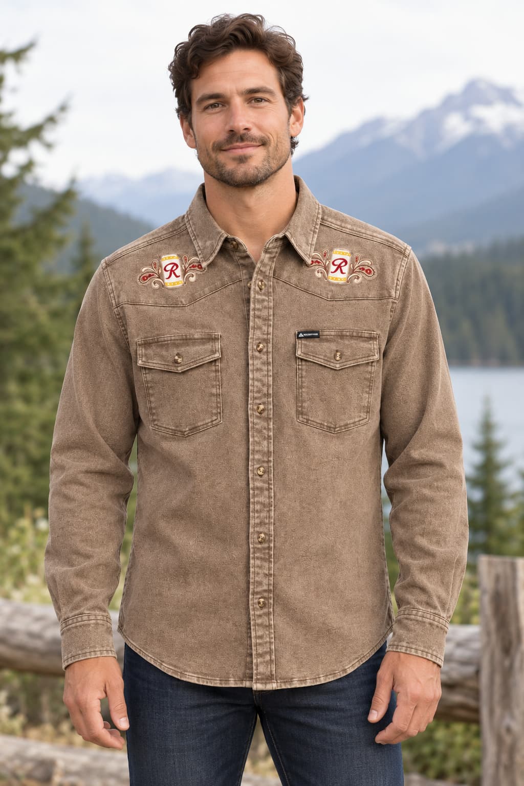 A man wearing a brown western-style button-up shirt with embroidered chest details, standing outdoors in front of mountains and a lake.