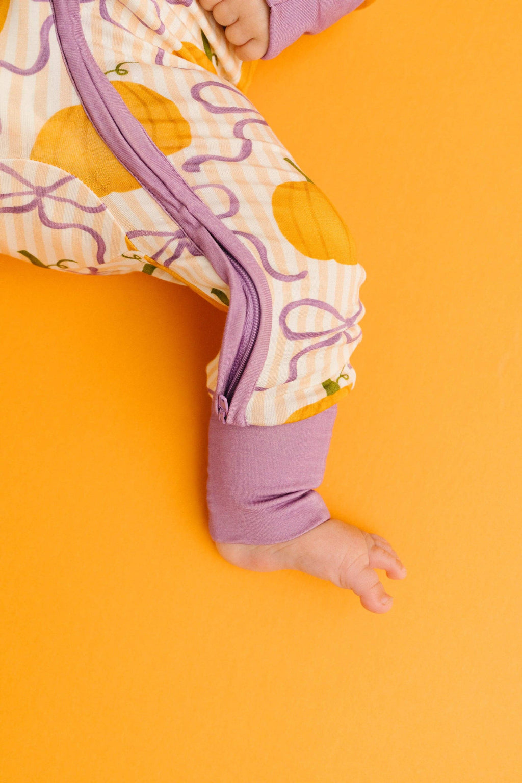 A baby's foot and lower leg wearing a patterned footless sleeper with purple cuffs, laid on a solid orange background.
