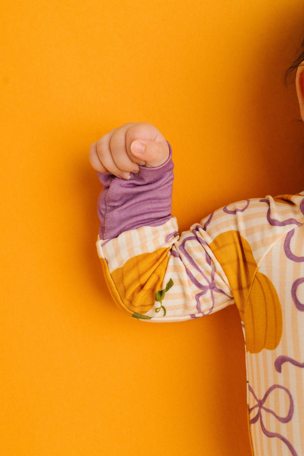 A close-up of a baby's arm and hand wearing a purple long-sleeve shirt under a cream and yellow gingham bodysuit with a pumpkin print, posed against a solid orange background.