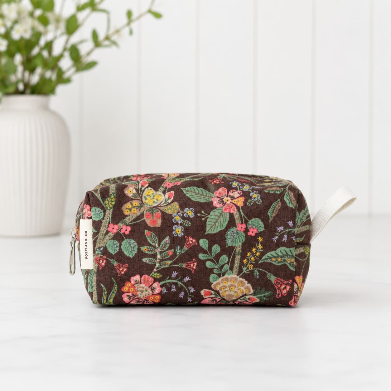A rectangular cosmetic pouch with a brown background and a colorful floral print, featuring a white fabric loop handle and a small white brand tag on the side, set against a white paneled background with a vase of flowers.