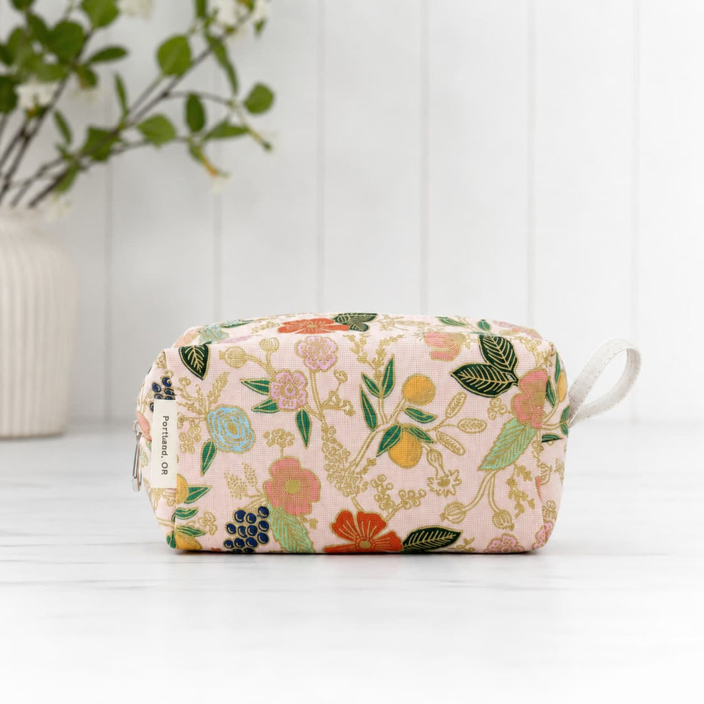 A rectangular zippered pouch with a light pink background and a colorful floral print, featuring a white fabric loop handle on the right side. The brand name 'KIMONO' is visible on a small fabric tag on the left side.