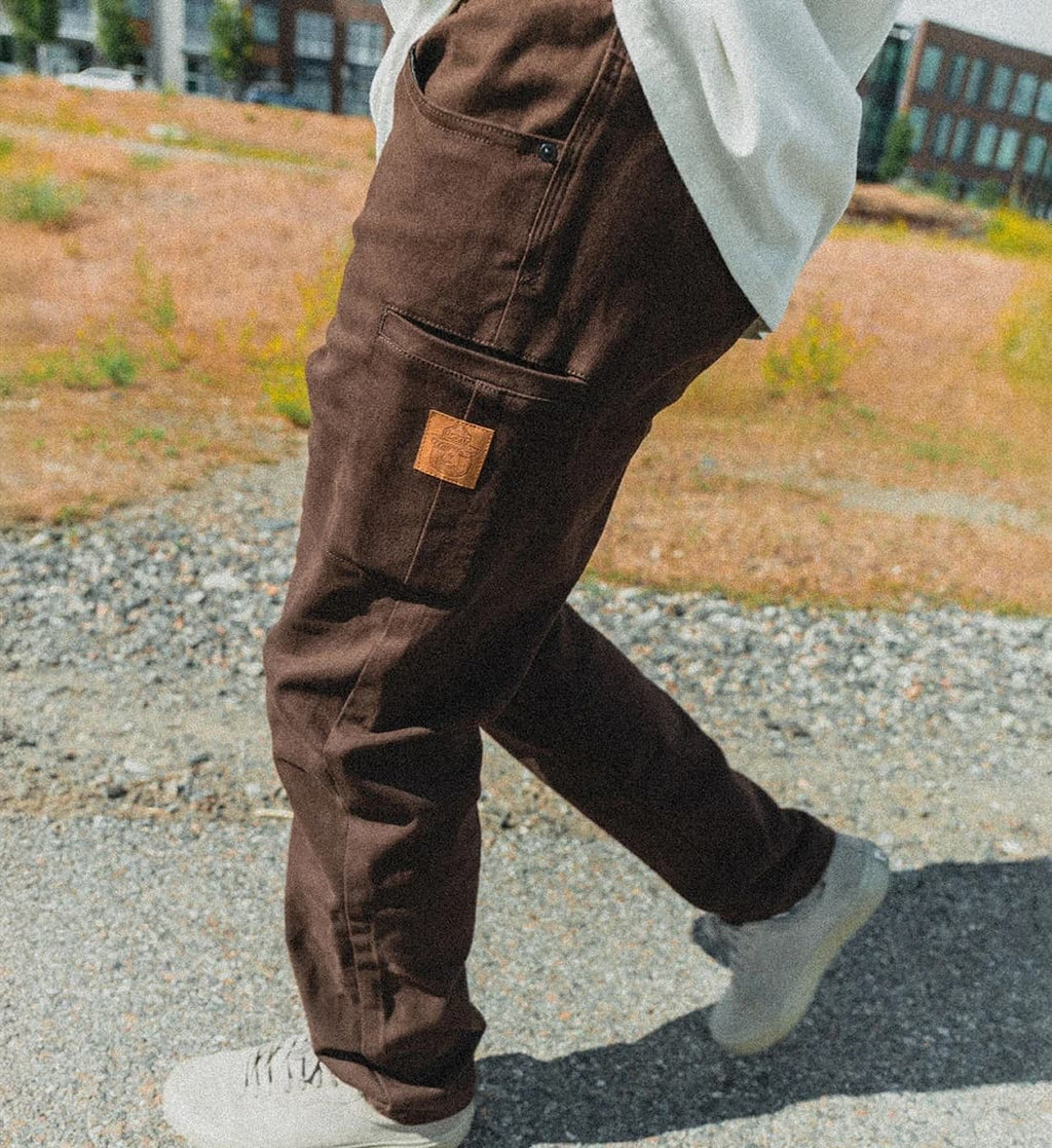 A person wearing dark brown canvas cargo pants with a small brown leather patch on the pocket, walking outdoors on a gravel path.