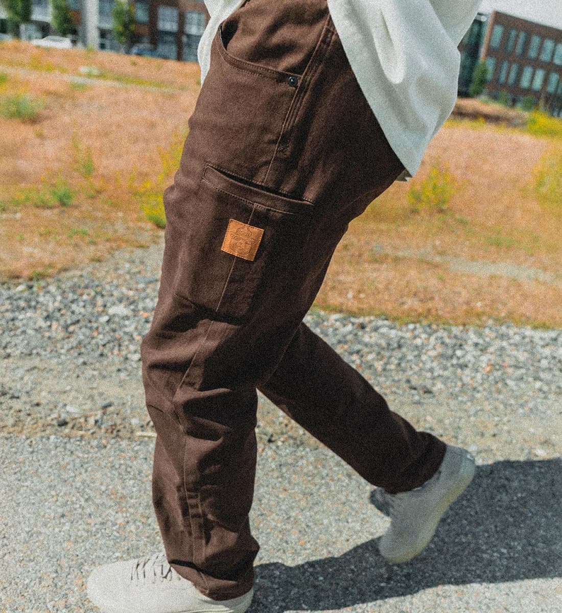 A person wearing dark brown canvas cargo pants with a small brown leather patch on the pocket, walking outdoors on a gravel path.
