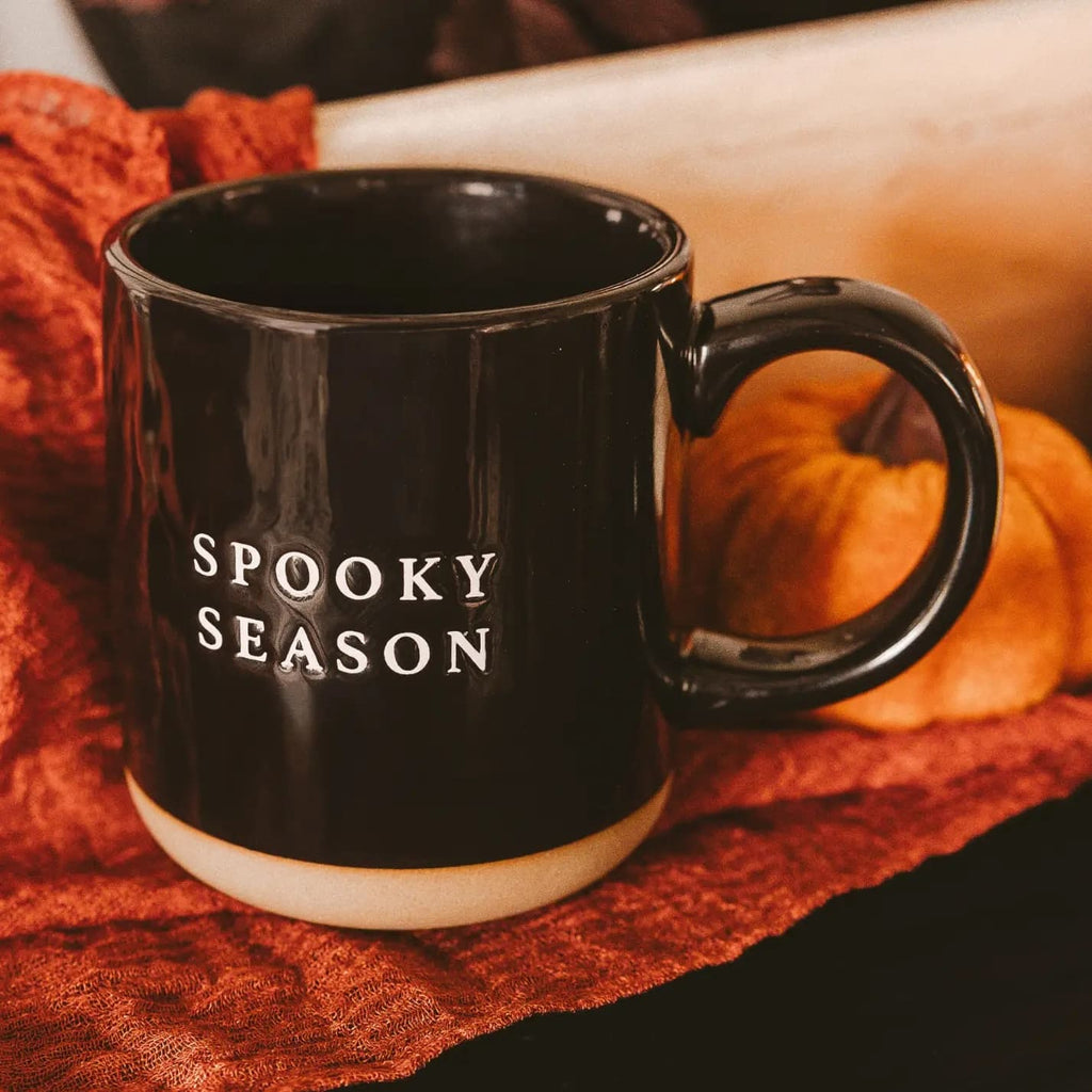 A glossy black ceramic mug with a cream-colored base and the words 'SPOOKY SEASON' printed in white on the side, styled on a textured orange fabric with autumn props in the background.