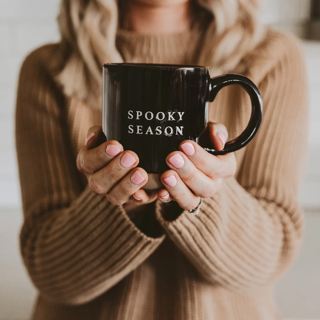 Person holding a black mug with 'SPOOKY SEASON' text, wearing a beige sweater.