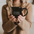 Person holding a black mug with 'SPOOKY SEASON' text, wearing a beige sweater.