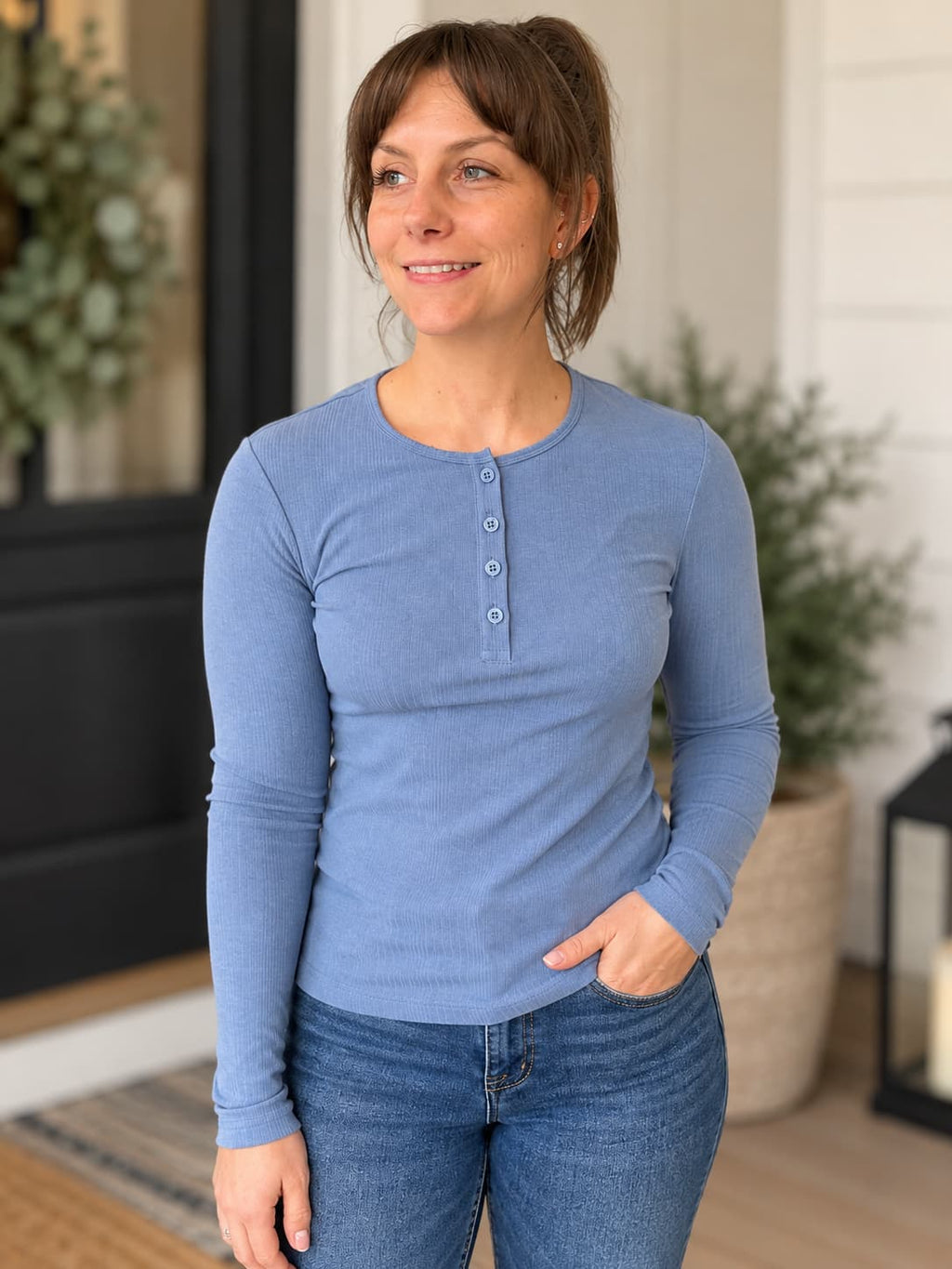A woman wearing a light blue long-sleeve henley shirt and blue jeans, standing on a porch with potted plants in the background.