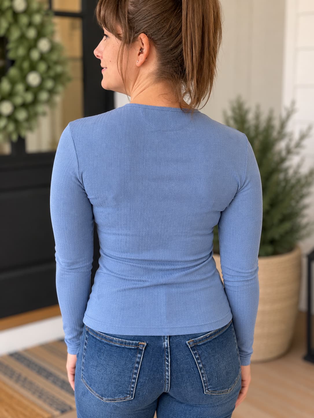 A woman shown from the back wearing a fitted long-sleeve ribbed top in dusty light blue, paired with blue jeans, standing indoors with potted plants in the background.