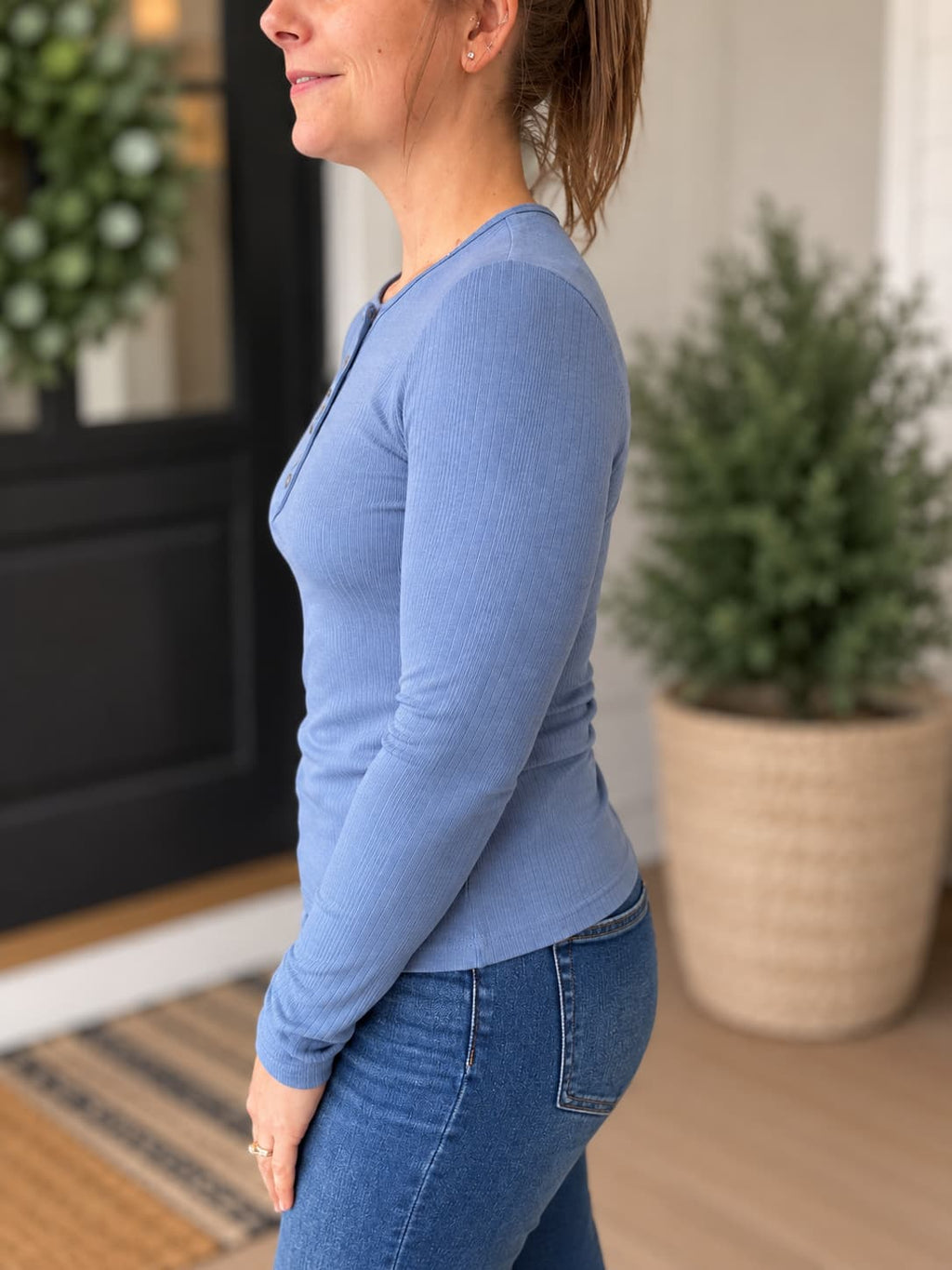 A woman shown in side profile wearing a fitted long-sleeve knit top in dusty light blue with blue jeans, standing indoors near a door and potted plants.