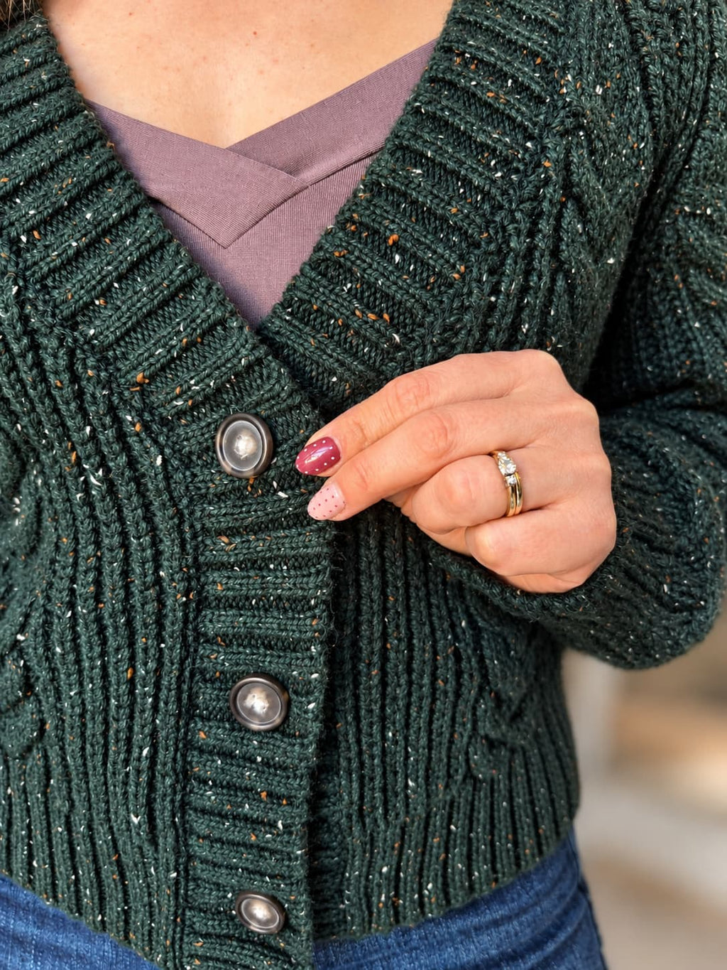 Close-up of a person wearing a dark green cable-knit cardigan with silver buttons over a mauve V-neck top and blue jeans. The cardigan has subtle orange and white flecks throughout.